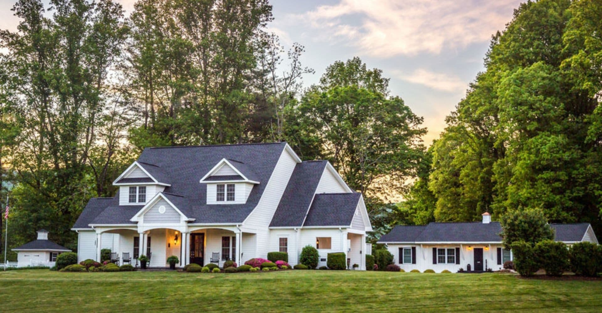 A large, white colonial-style house with a black roof, surrounded by lush green trees and a manicured lawn.