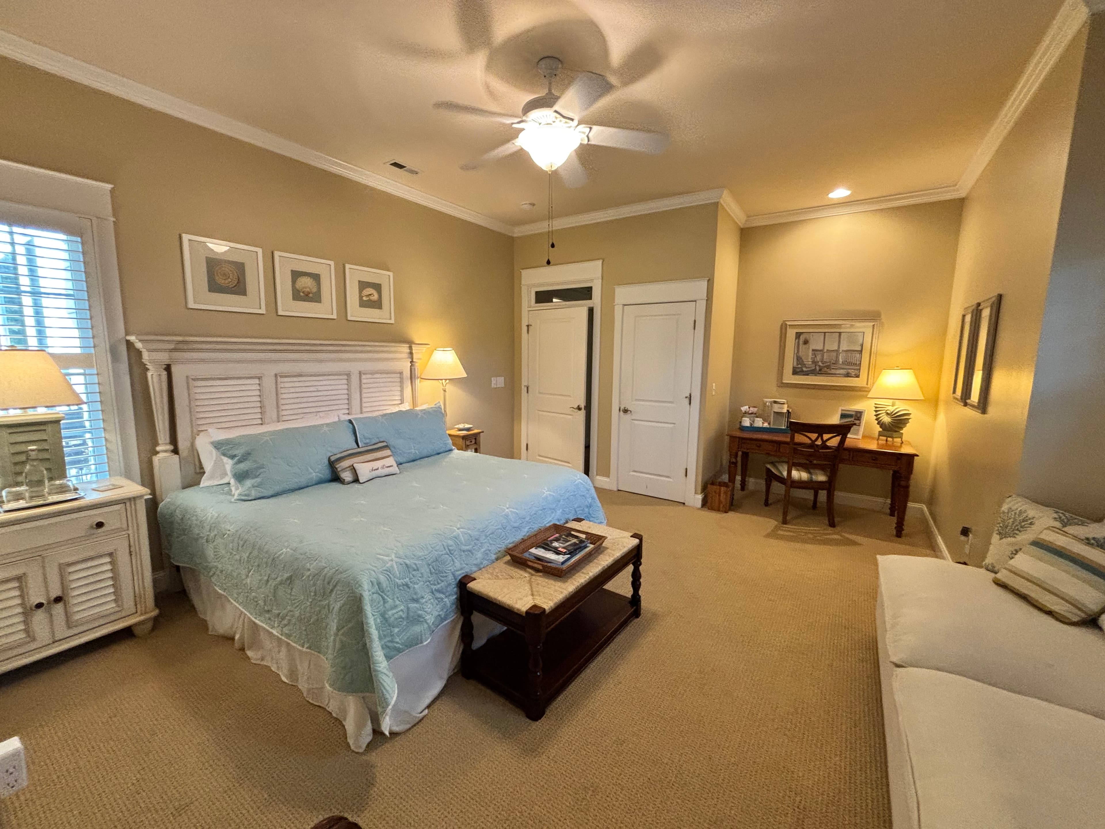 "Elegant guest room with king bed, light blue bedding, and beach-inspired décor. Features a writing desk with coffee station, cozy sitting area, and warm ambient lighting - perfect for a relaxing coastal getaway or bed and breakfast stay.