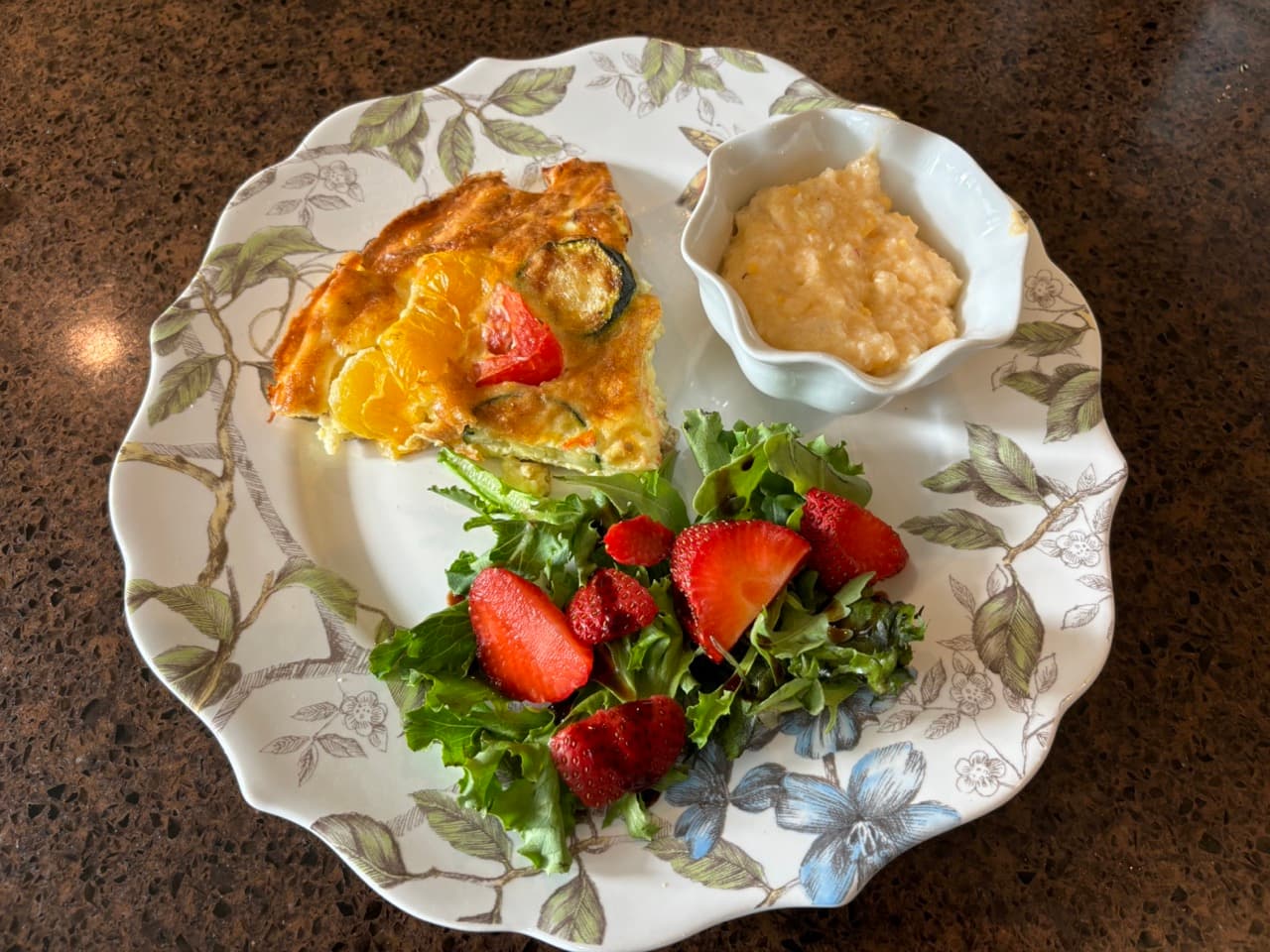 A plate featuring a slice of vegetable frittata, a side salad with strawberries, and a small bowl of grits.