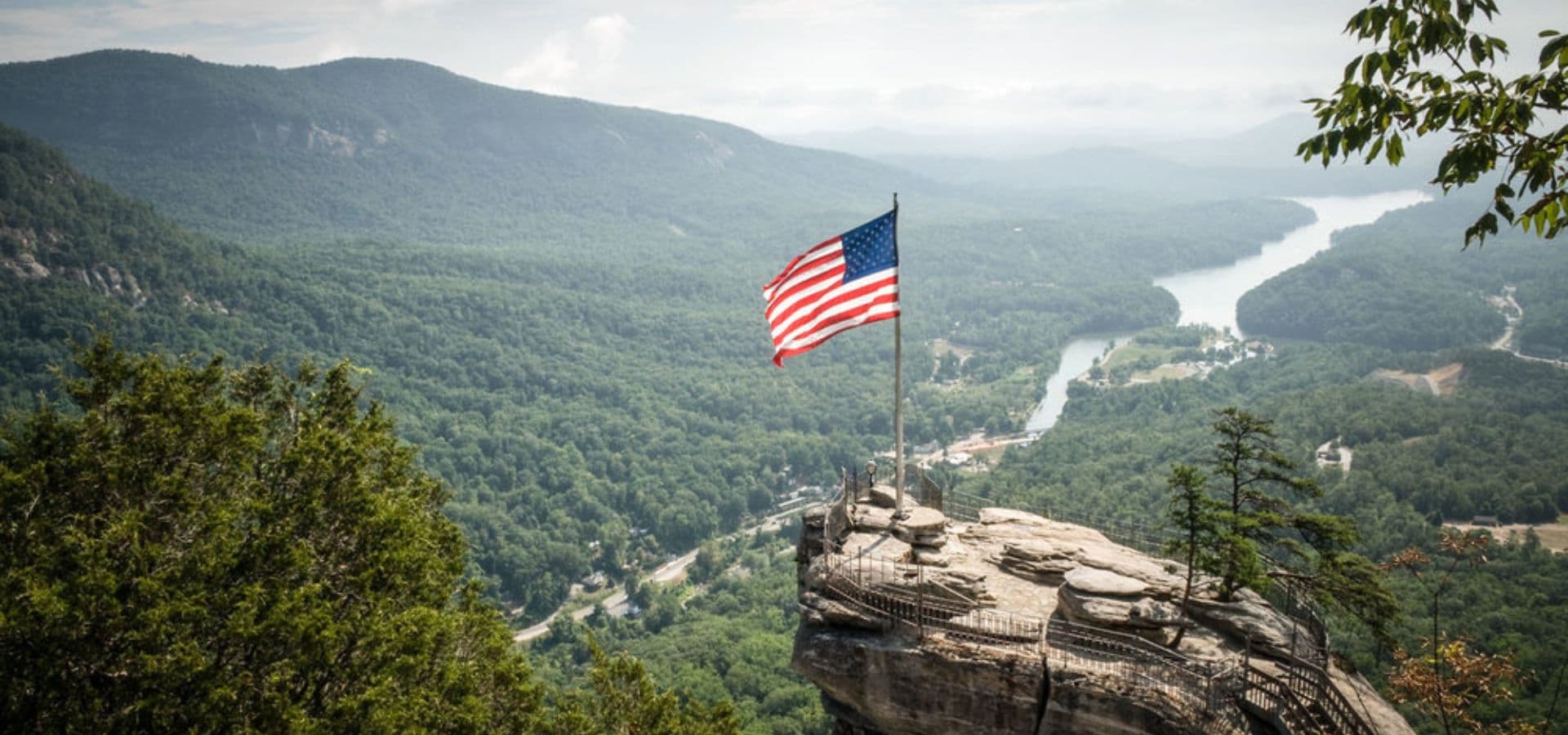 A flag stands atop a rocky outcrop overlooking a lush green valley and winding river.