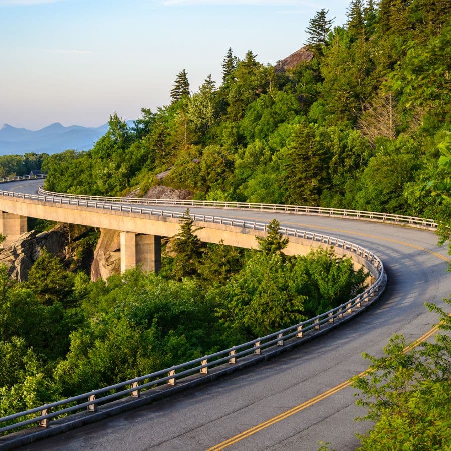 A winding road curves through lush greenery with a bridge spanning a rocky area.