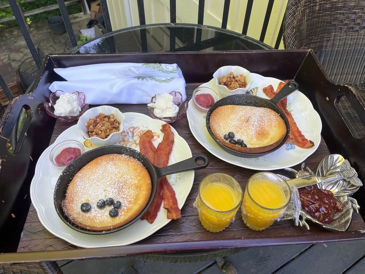 A breakfast tray featuring fluffy pancakes in cast iron skillets, crispy bacon, granola, whipped cream, jam, and two glasses of orange juice.