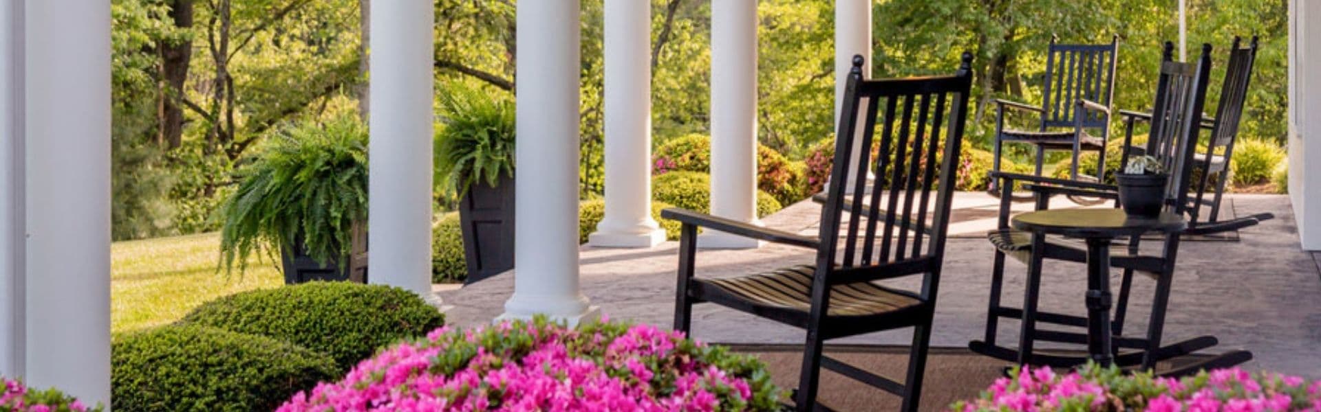 A serene porch adorned with pink flowers, rocking chairs, and lush greenery.