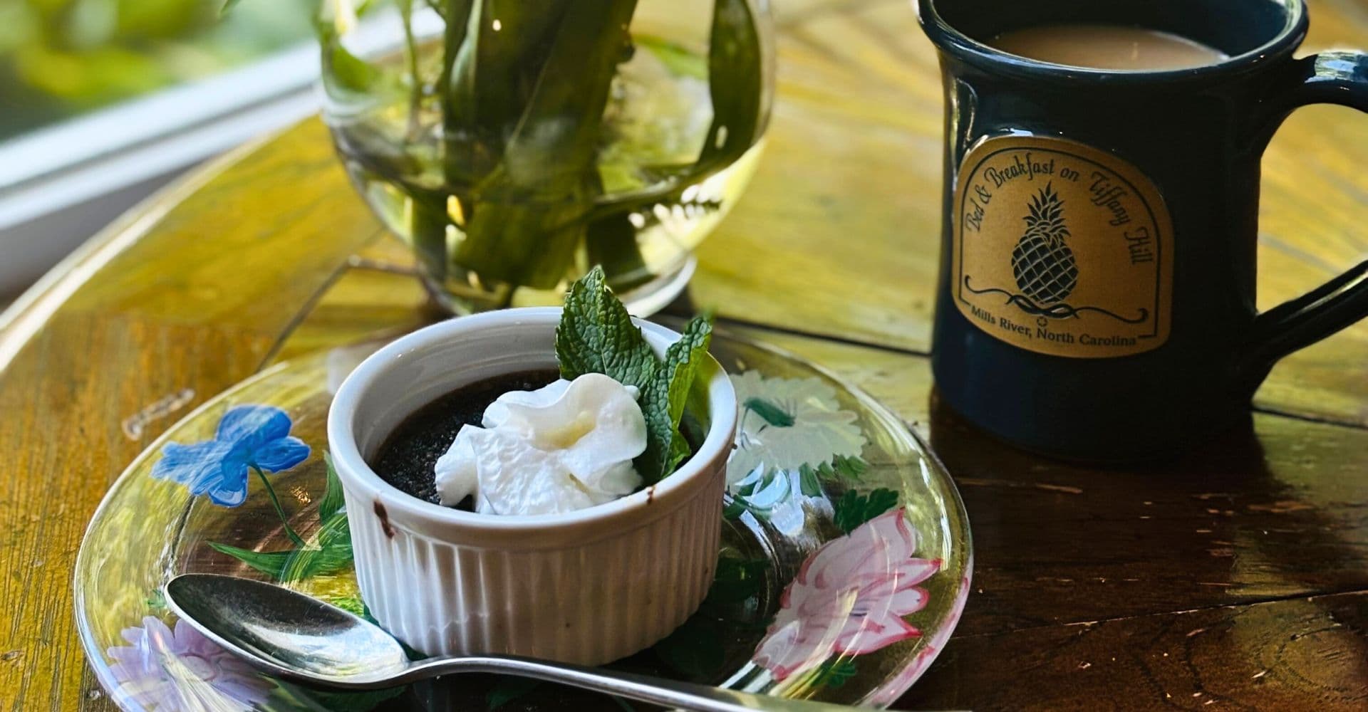 A chocolate dessert topped with whipped cream and mint, served with a mug of coffee on a floral plate.