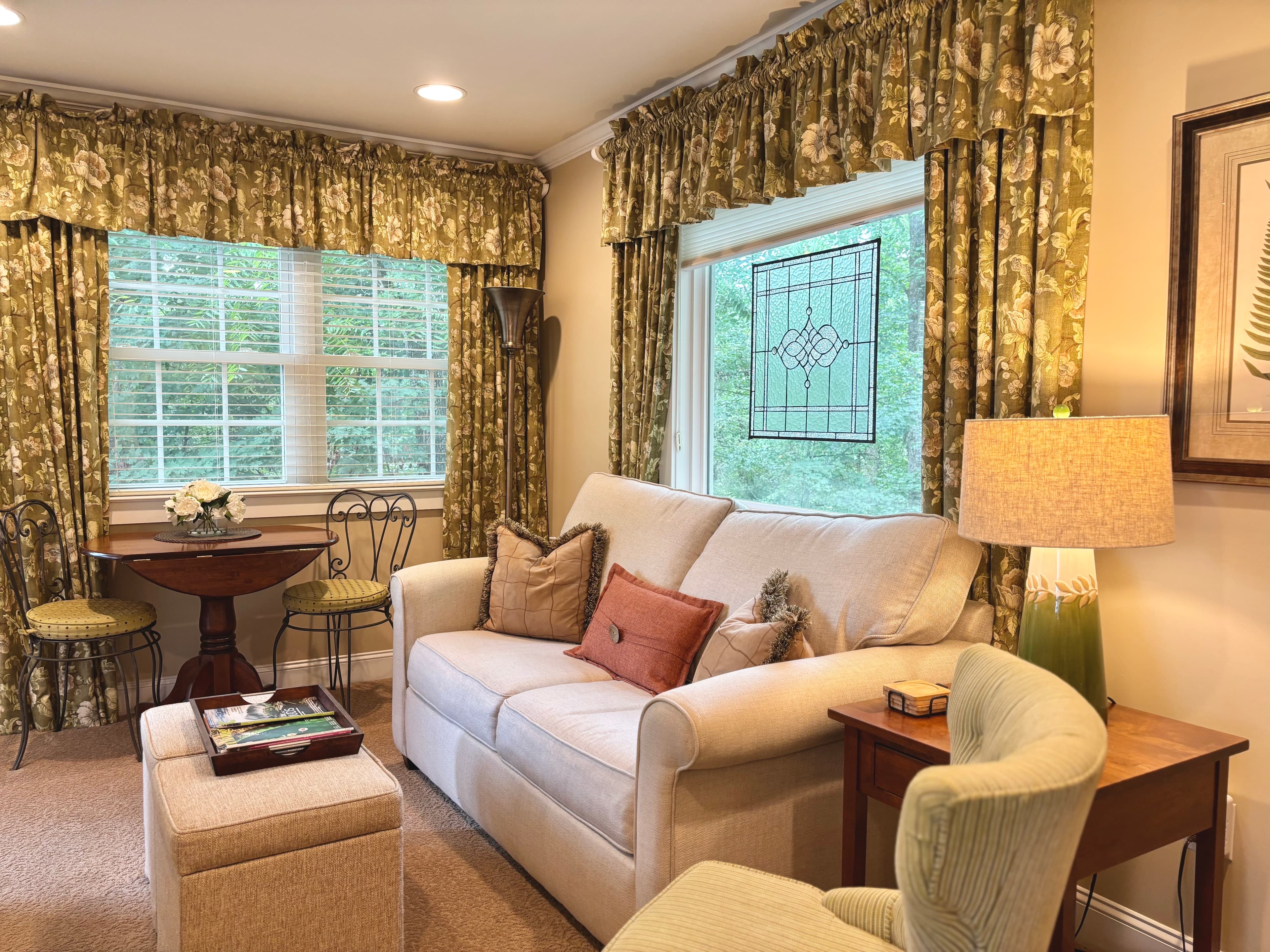 Cozy living room with a light-colored sofa, decorative curtains, and a small dining table by a window.