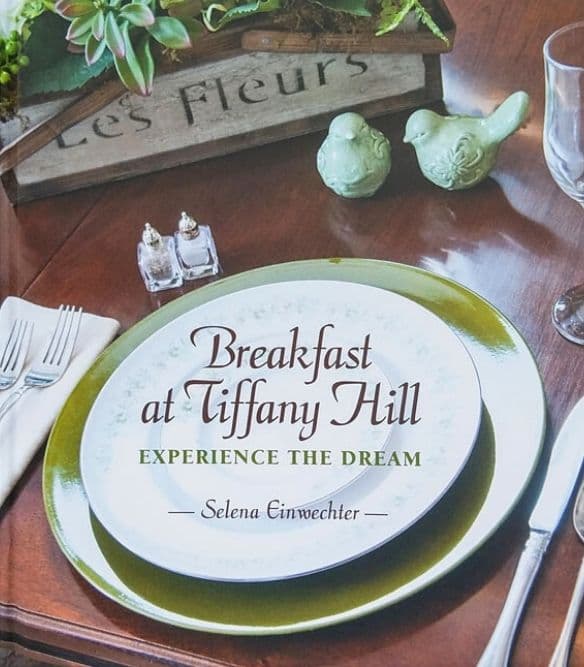 A table setting featuring a decorative plate, silverware, and two green bird figurines, with a book cover titled "Breakfast at Tiffany Hill."