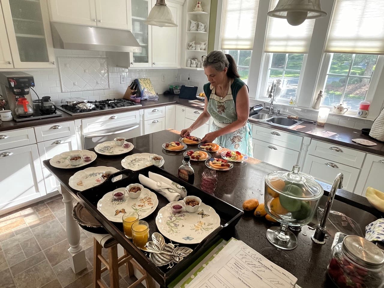 A woman in an apron prepares plates of fresh fruit in a bright kitchen.