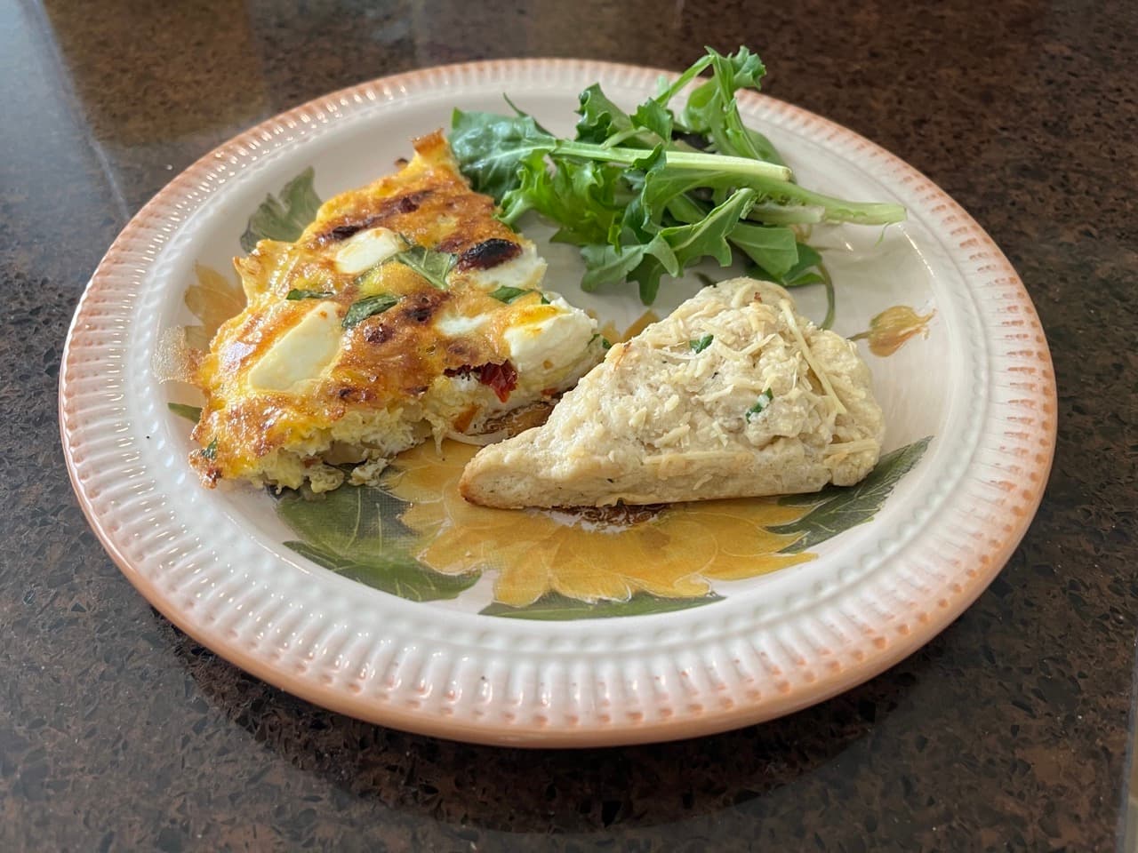 A plate featuring a slice of frittata, a scone, and a small serving of arugula.