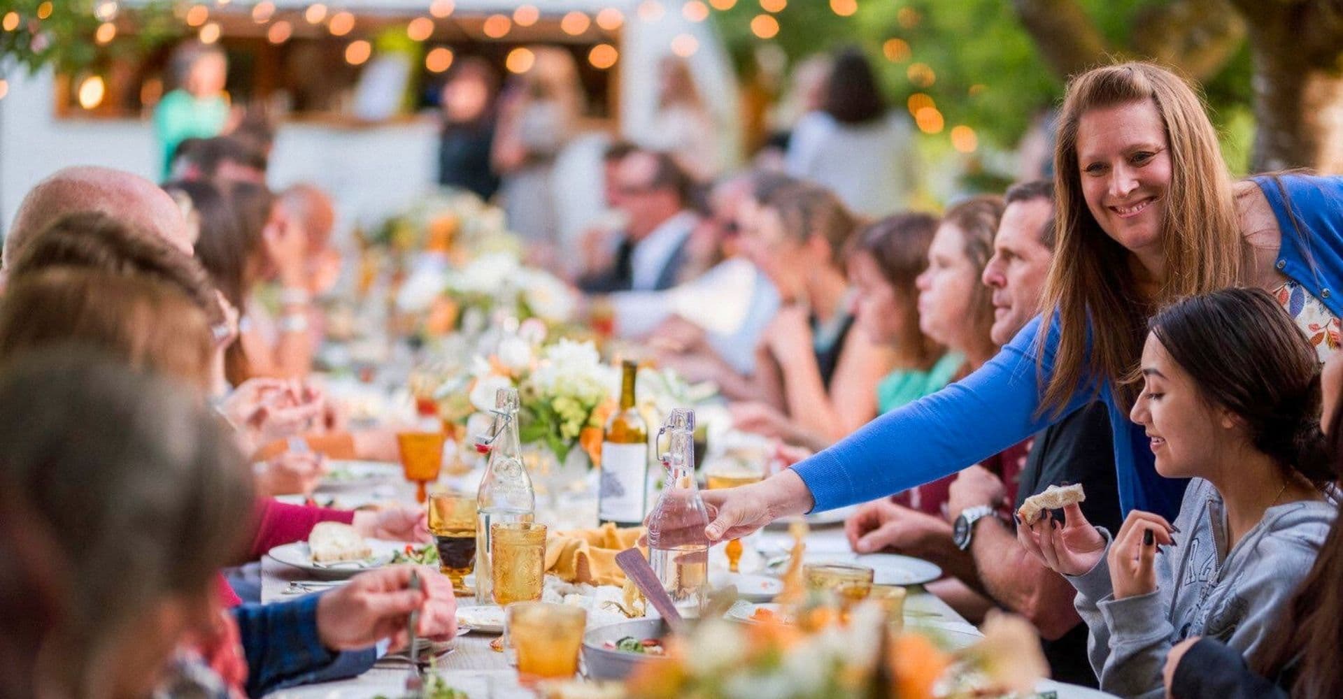 A woman serves water at a festive outdoor dining table filled with guests.