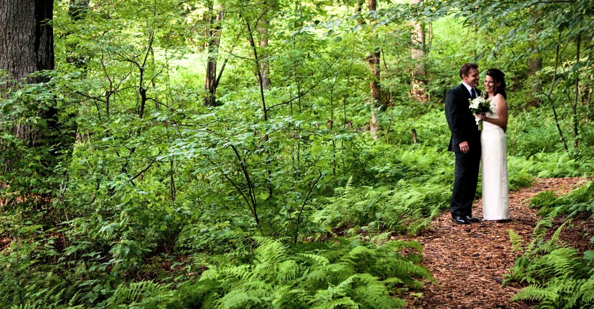 A bride and groom stand smiling together on a forest path surrounded by lush greenery.