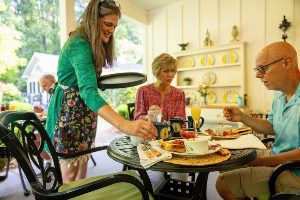 A woman serves food to two seated guests at a breakfast table on a porch.