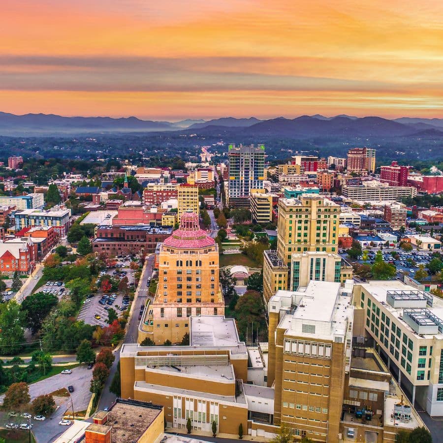 Aerial view of a city at sunset, showcasing a mix of modern and historic buildings against a mountain backdrop.