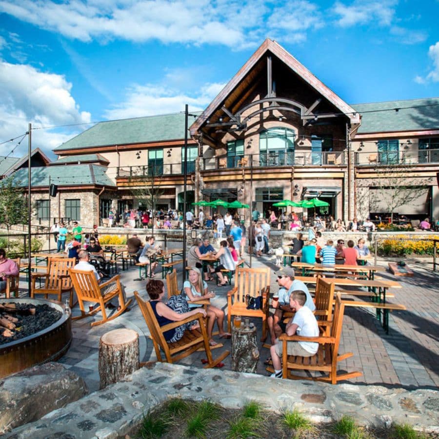 A busy outdoor gathering space with people seated at tables and rocking chairs near a building with large windows.