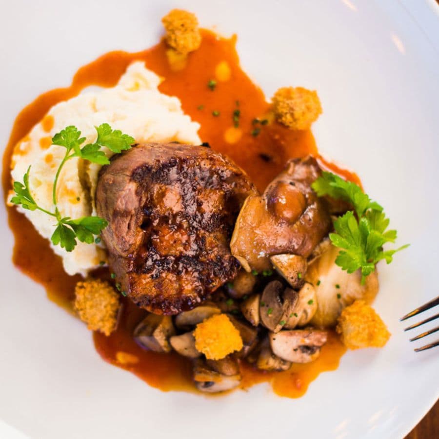 A beautifully plated dish featuring tender meat with mashed potatoes, sautéed mushrooms, and garnished with fresh herbs.