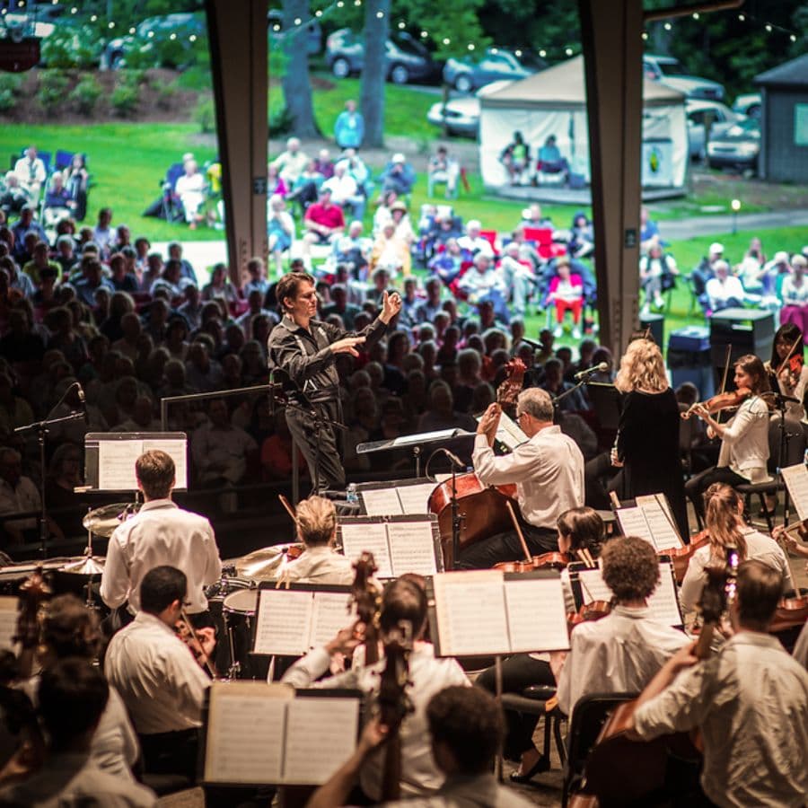 An orchestra performs on stage as a conductor leads the ensemble, with a large audience seated in the background.
